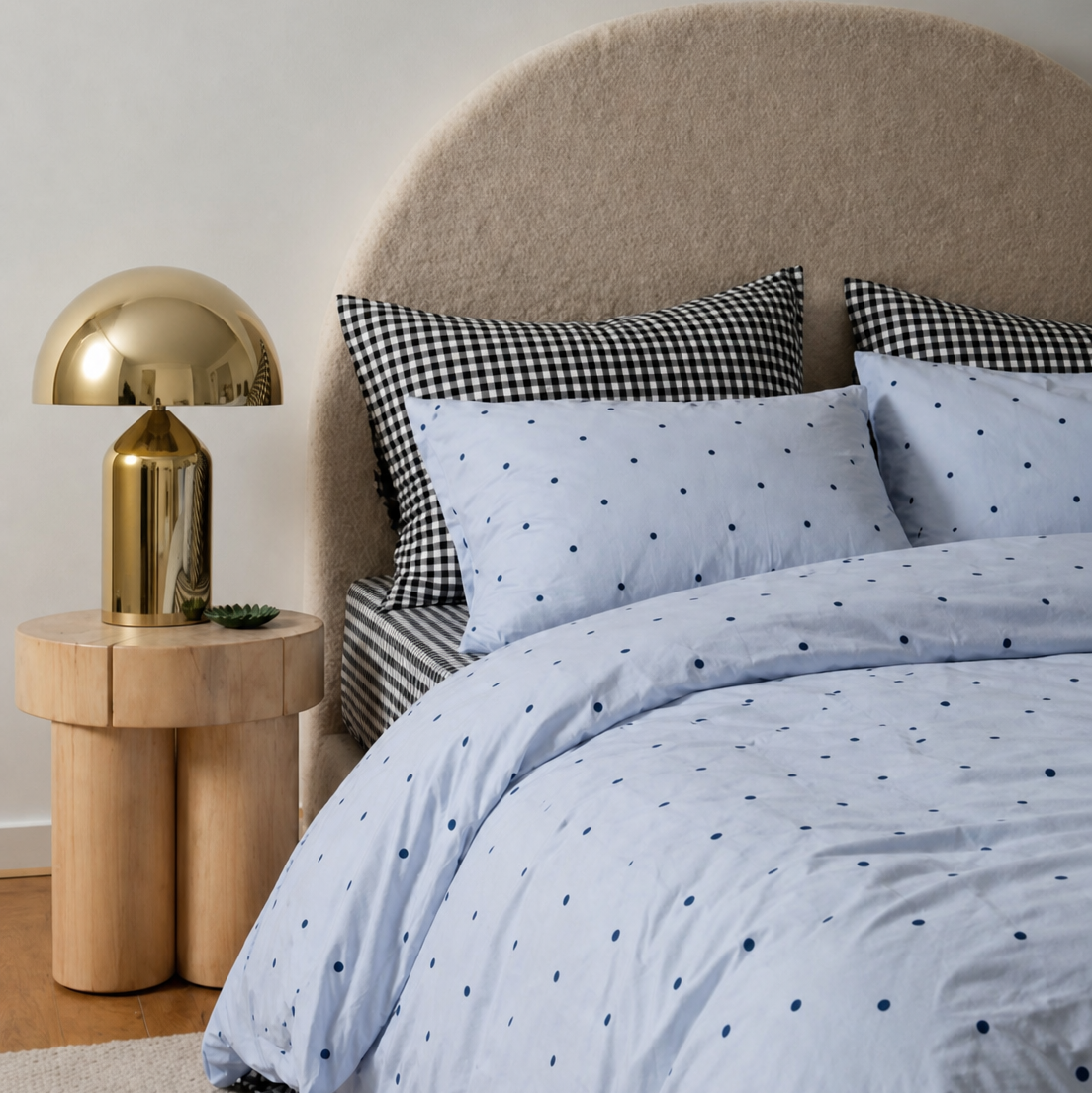 Bed with blue polka dot bedding, checkered pillows, and a gold lamp on a wooden side table.