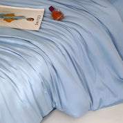 Close-up of pale blue Agnes Silk Bedding Set, soft sheen and draped folds, styled with magazine and perfume bottle