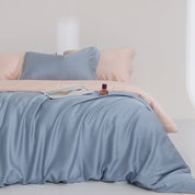 Agnes Silk Bedding Set in Sky Blue, draped over peach sheets with a magazine and perfume styled on top