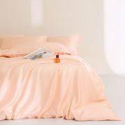 Agnes Silk Bedding Set in soft peach, made bed with silky sheen, styled with a magazine and perfume
