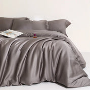 Agnes Silk Bedding Set in slate grey, glossy silk quilt cover draped with soft folds, magazine and perfume on bed