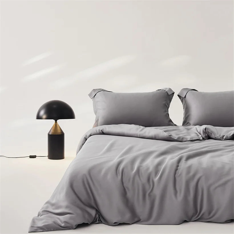 Amélie Bedding Set in Dove Grey, sheened quilt cover and pillowcases on a bed beside a black table lamp