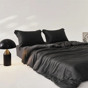 Amélie Bedding Set in Noir, deep black lyocell silk with soft sheen, styled on a bed beside a dome lamp
