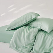 Amélie Bedding Set in soft sage green, pillowcases piled on a rumpled quilt cover showing the silky sheen