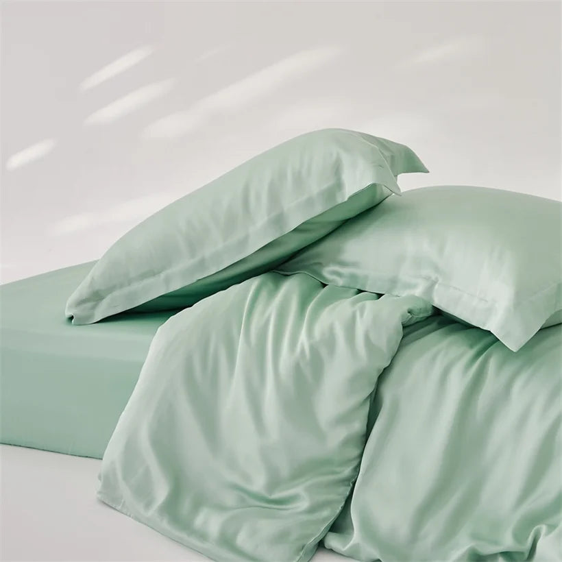 Amélie Bedding Set in soft sage green, pillowcases piled on a rumpled quilt cover showing the silky sheen