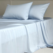 Amélie Flat Sheet Set in Celeste Blue, draped over a bed with two pillowcases showing a soft sheen