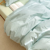 Close view of the Blossom Bow Bedding Set in Mint, fine green and white stripes draped over a bed with an open book