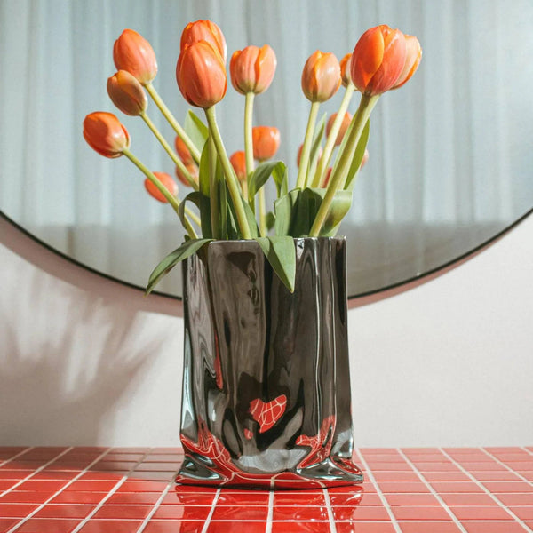 Chrome Vase in mirrored gunmetal holds orange tulips on a red tiled table before a round mirror
