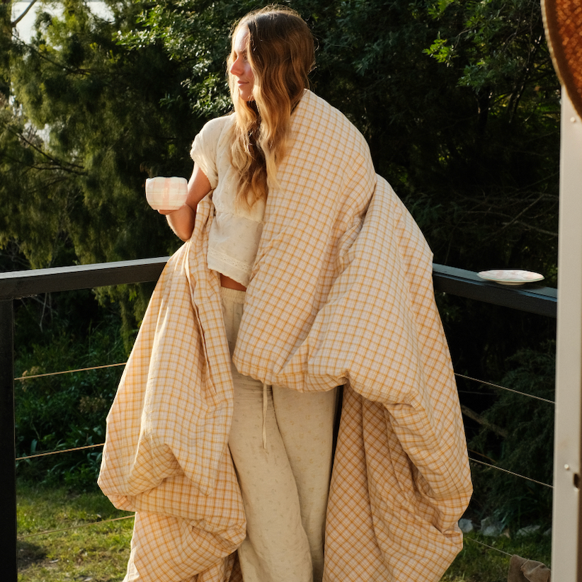 Woman wrapped in the Dakota Bedding Set quilt cover, golden plaid on cream, holding a mug outdoors
