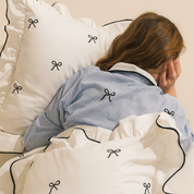 Isabella Bow Bedding Set in ivory with black bow embroidery and piping, model resting against pillows