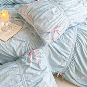 Metaya Bedding Set in Sky Blue, ruched corners tied with pink ribbon, candle and book beside the bed