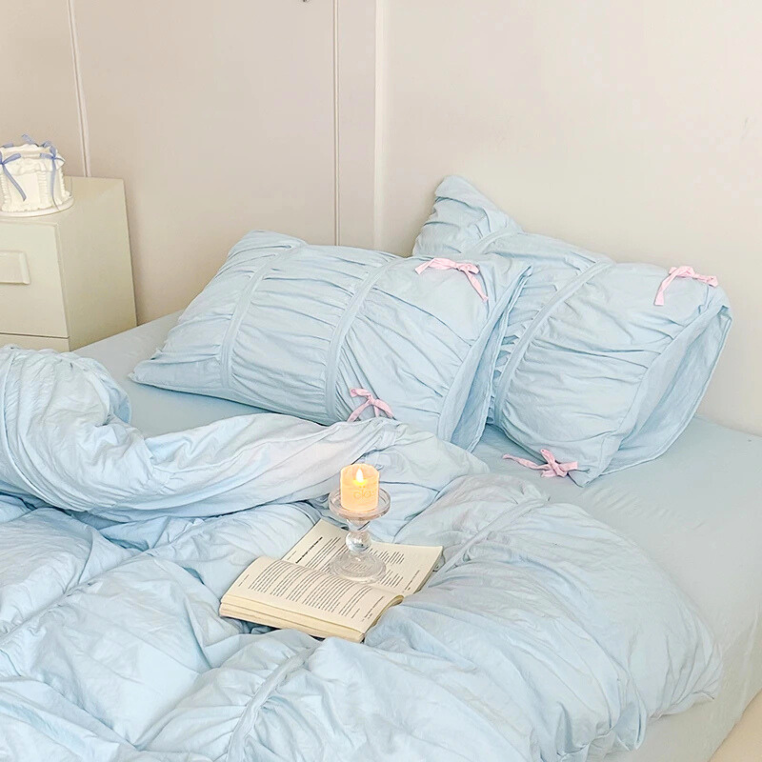 Metaya Bedding Set in Sky Blue, ruched pillowcases tied with pink bows, book and candle on the bed
