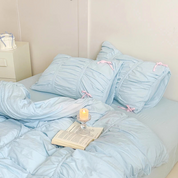 Metaya Bedding Set in Sky Blue, ruched pillowcases tied with pink bows, book and candle on the bed