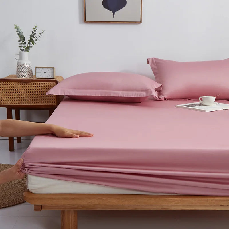 Hands fit the elasticated edge of the Sola Cotton Fitted Sheet in Blush pink onto a wooden bed frame