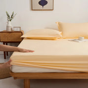 Hands tuck the Sola Cotton Fitted Sheet in Butter over a mattress corner, elasticated edge visible