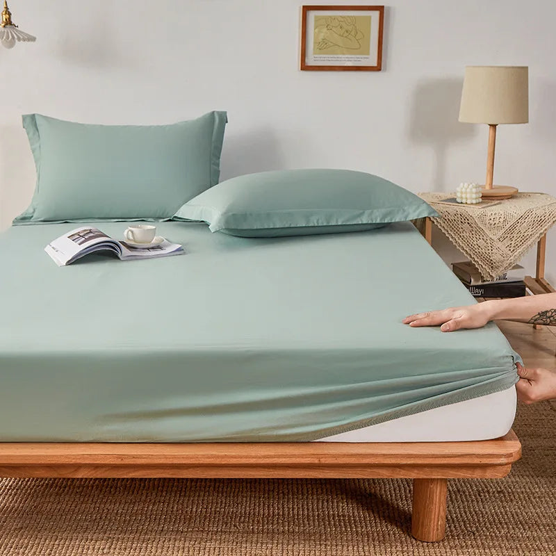 Hands tuck the elasticated corner of the Sola Cotton Fitted Sheet in sage green onto a wooden bed frame