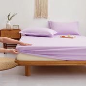 Hands pull the elasticated corner of the Sola Cotton Fitted Sheet in Lilac onto a wooden bed frame