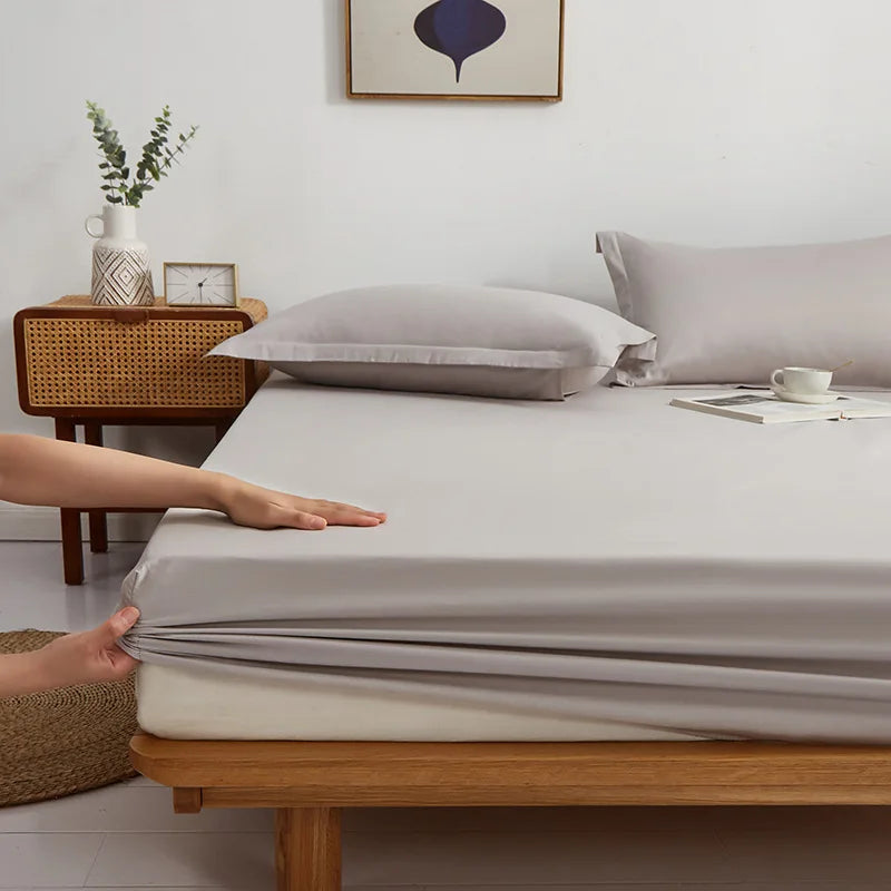 Hands pull the elasticated corner of the Sola Cotton Fitted Sheet in soft grey onto a mattress