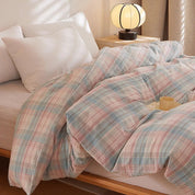 Soleil Stripe Bedding Set in soft pink and blue check weave, rumpled on a bed beside a lamp and mug