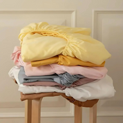Tate Fitted Sheet in soft yellow, folded atop a stack of mixed-colour sheets on a wooden stool