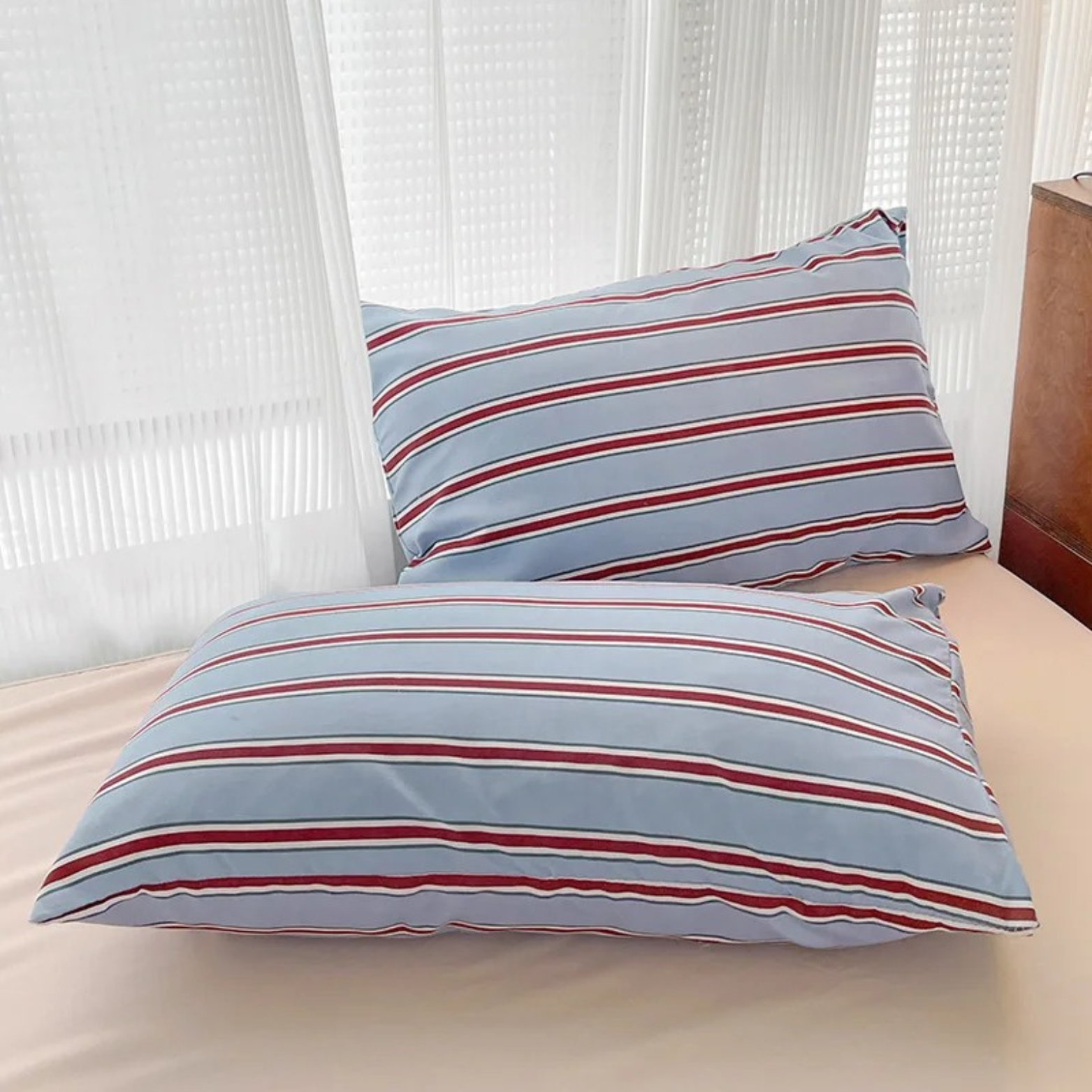Two pillowcases from the Vida Bedding Set in Blue & Red, striped, propped against a bed by sheer curtains
