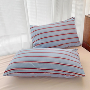 Two pillowcases from the Vida Bedding Set in Blue & Red, striped, propped against a bed by sheer curtains