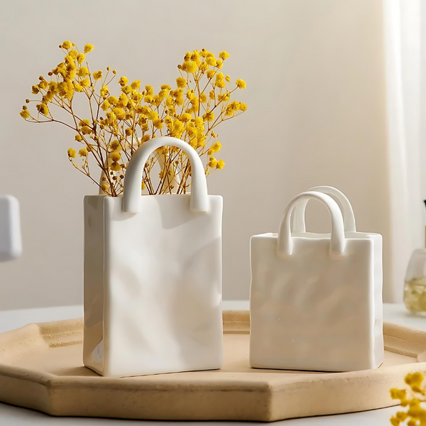 Two sizes of the Vivien Handbag Vase in matte white, one holding yellow dried flowers, on a wooden tray